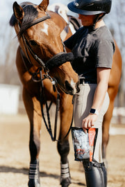 Rider standing beside her horse holding a bag of Enjoy Yums carrot horse treats – all-natural, baked heart-shaped biscuits made with real carrots.